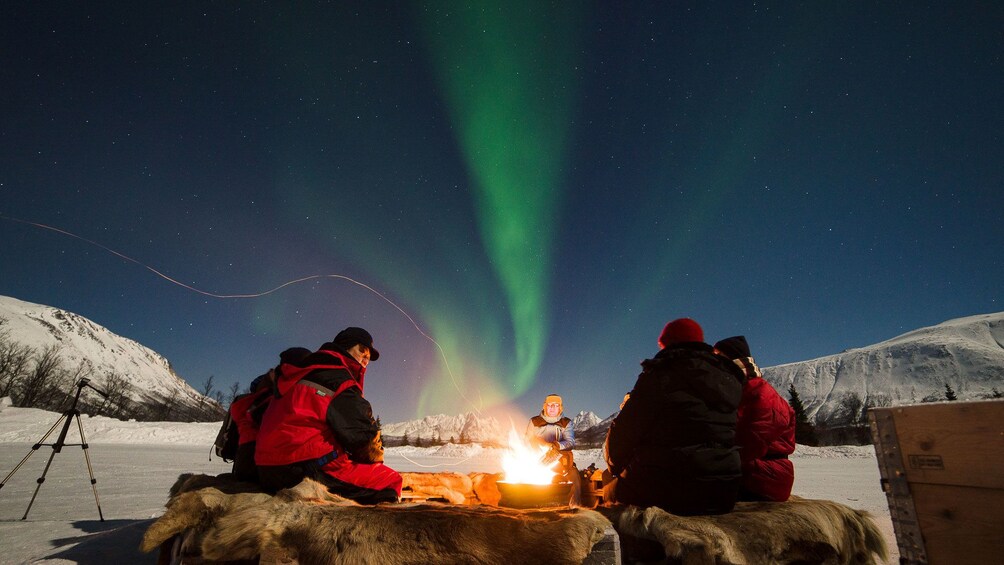 Adventurers sit around fire as Aurora Borealis streaks above in Tromso