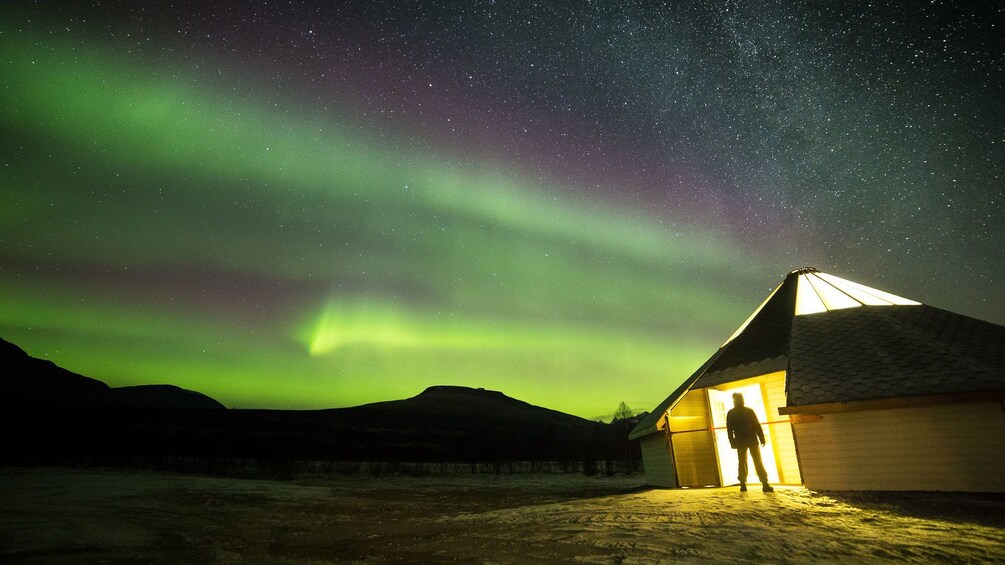 Man stands at entrance of hut with Aurora Borealis overhead in Tromso