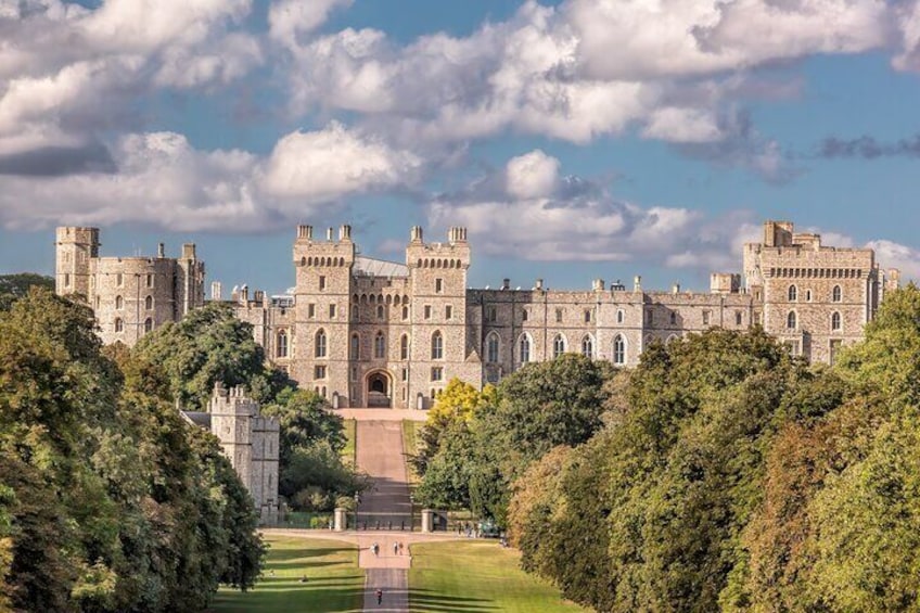 Windsor Castle seen from the Long Walk, framed by trees, highlighting the majestic structure standing proudly against a backdrop of clouds.