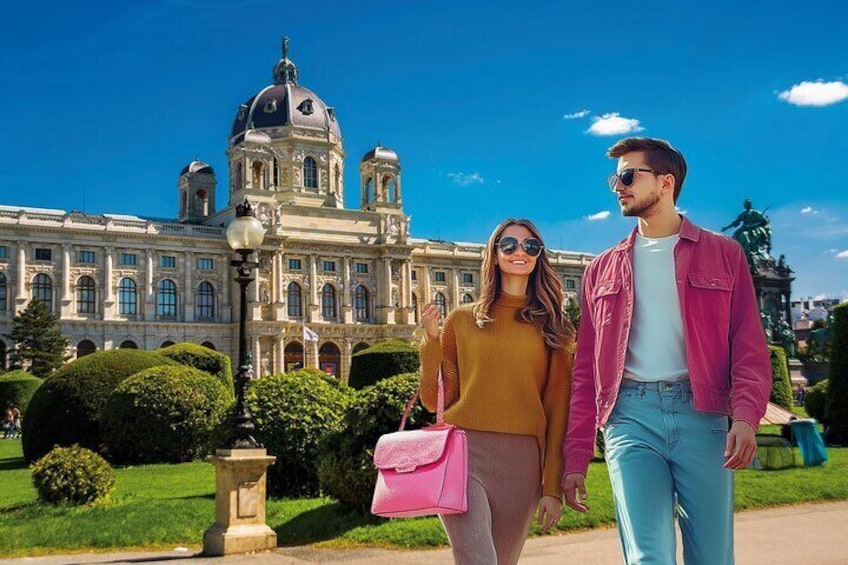 Stylish couple wearing sunglasses walk in front of the Kunsthistorisches Museum Vienna under bright blue skies.