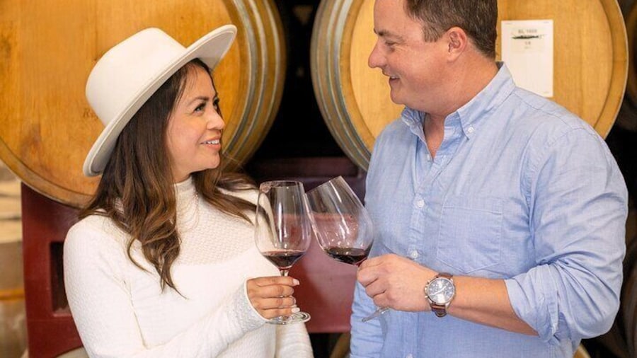 A couple enjoying a wine tasting inside a Santa Ynez Valley winery, toasting with red wine in front of stacked oak barrels during the Santa Ynez Full-Day Wine Experience with Picnic Lunch.