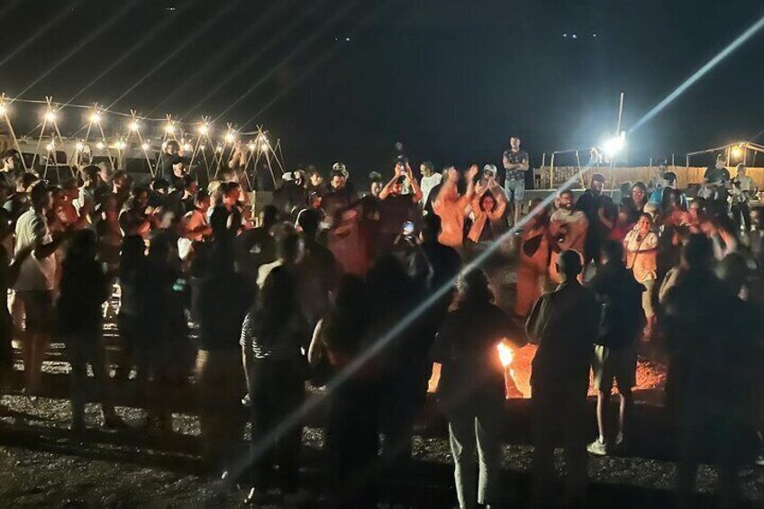 Feel the energy and joy of a lively gathering in the Agafay Desert. Under a canopy of stars, a spirited group dances and claps around a glowing campfire, creating an unforgettable ambiance of warmth a