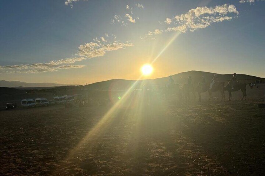 As the golden sun dips below the horizon, the Agafay Desert comes alive with breathtaking views and tranquil moments. In the foreground, silhouetted camel riders traverse the sandy terrain, creating a
