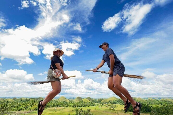 Two tourists humorously pose as if flying on broomsticks over the Chocolate Hills, with a bright blue sky and green hills below.