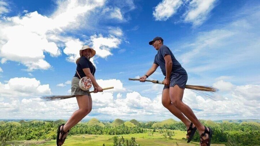 Two tourists humorously pose as if flying on broomsticks over the Chocolate Hills, with a bright blue sky and green hills below.