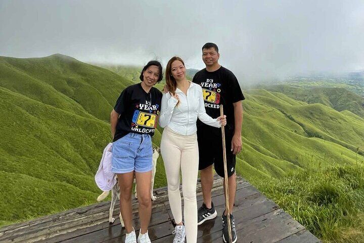 Three hikers pose on a wooden platform at Alicia Panoramic Park, surrounded by a stunning expanse of mist-covered rolling green hills.