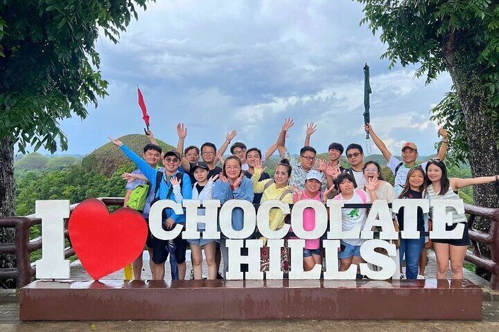 A cheerful group of tourists poses behind a large “I ❤️ Chocolate Hills” sign, with the iconic Bohol hills in the background.