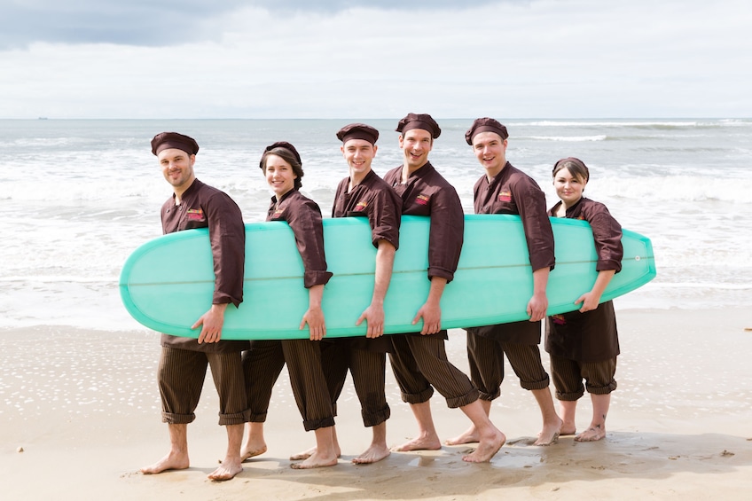 Six people hold a surf board on a beach