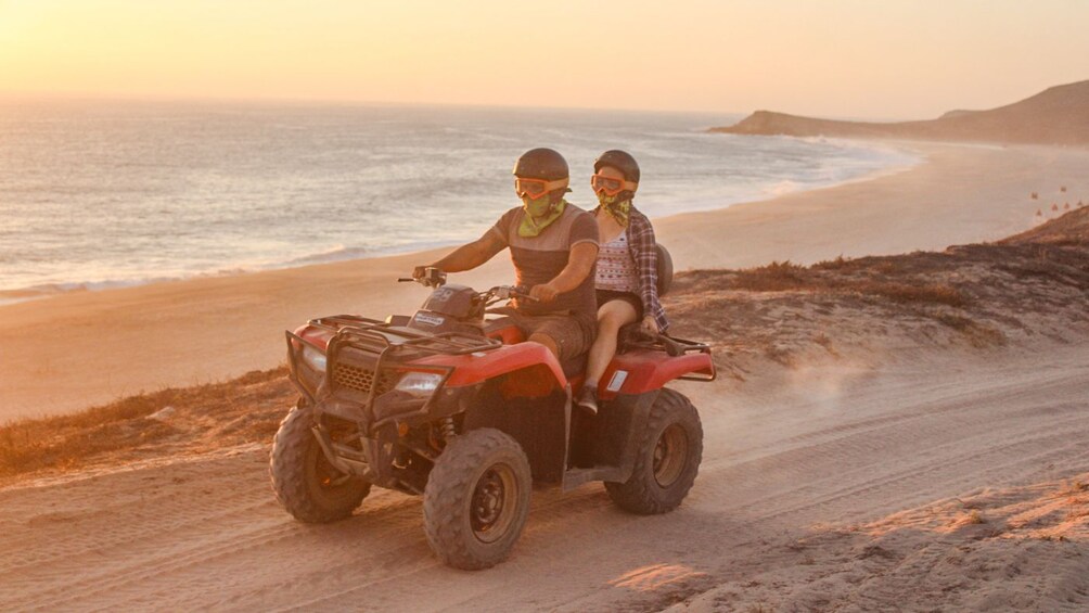 Two people ride an ATV on a dirt road next to the ocean at sunset