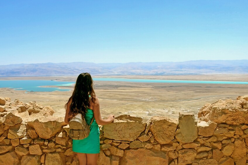 Woman stands next to a Rock wall and looks out at the Dead Sea