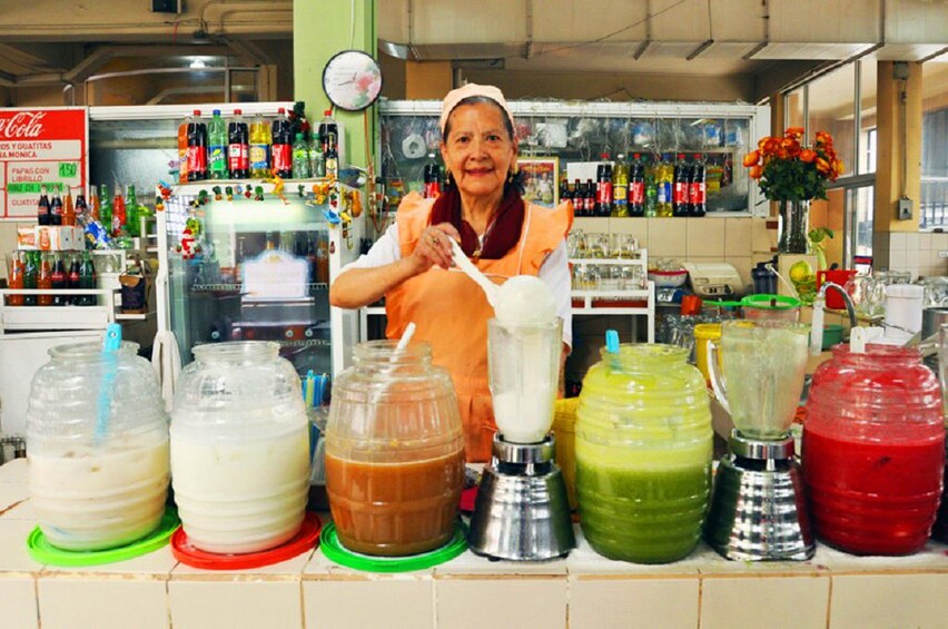Woman stands next to large containers of multicolored juices at a restaurant in Quito