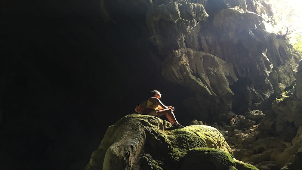 Woman sits on large rock near sunny cave opening of Tham Chang Cave