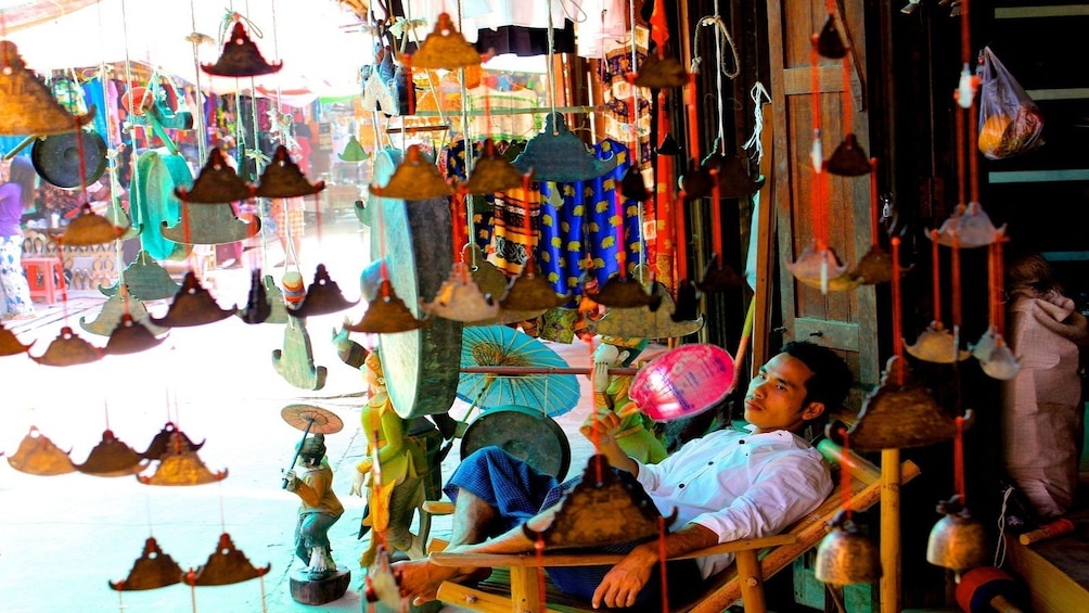 Man relaxes on a chair holding a pink fan and surrounded by jewelry in Mani Sithu Market