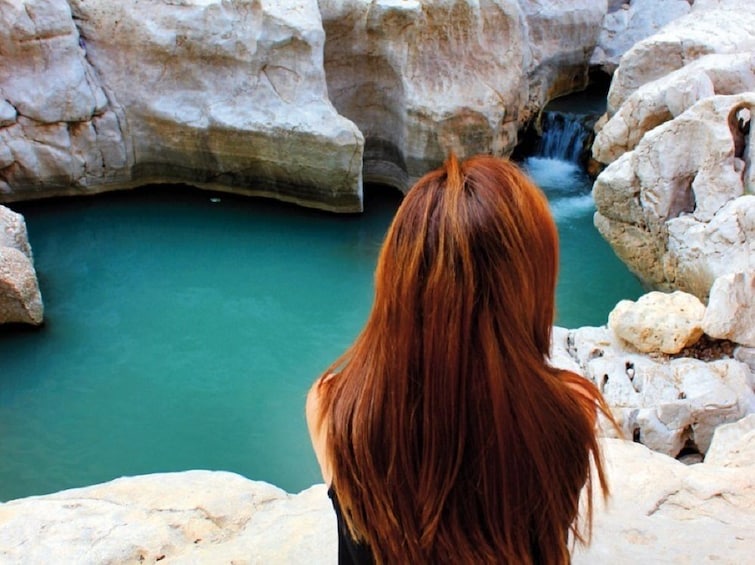 From behind, a woman looks over the clear pools at Wadi Ash