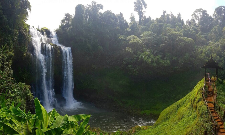 Panoramic view of Tad Yuang Waterfalls, lush surroundings and a walkway across the water