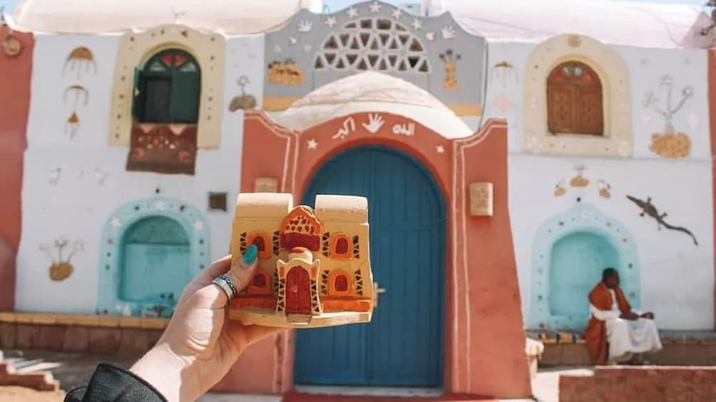 Hand holds small clay model of Nubian home with real home in the background