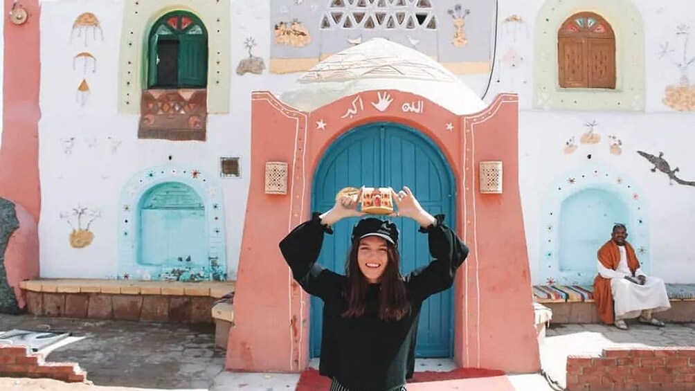 Woman with hat poses with small clay model over her head in front of Nubian house