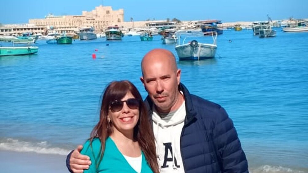 Couple pose in front of boats in the Mediterranean with Citadel of Qaitbay in the distance