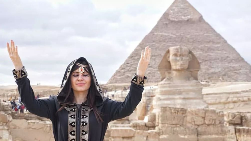 Woman poses with hands raised in front of sphinx and pyramid