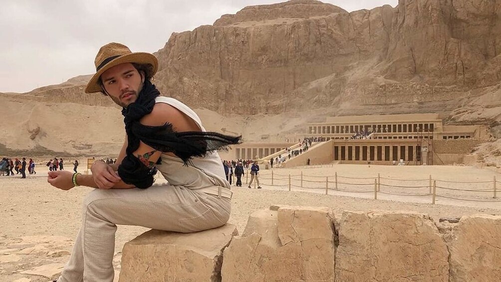 Man sits on short wall of stones with the Mortuary Temple of Hatshepsut in the background