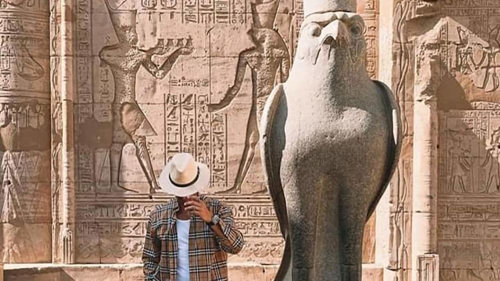 Man poses with hat covering his face next to statue of Horus at the Temple of Horus at Edfu