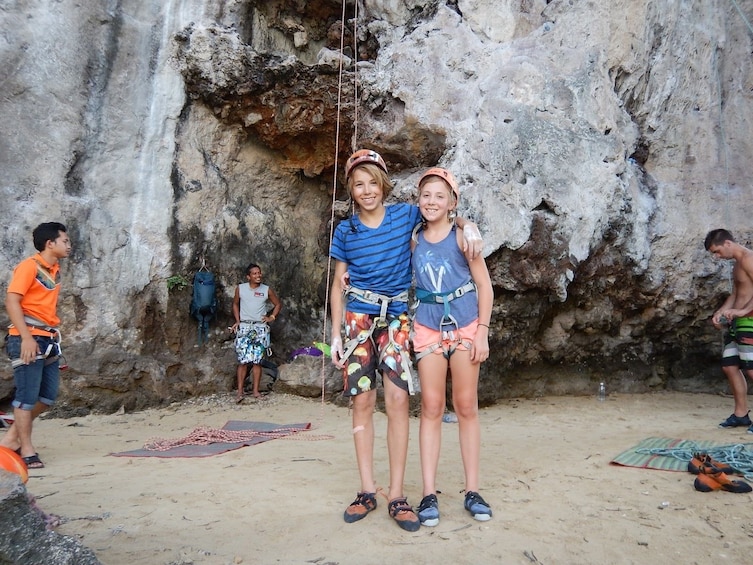 Two kids in harnesses pose in front of outdoor rock wall