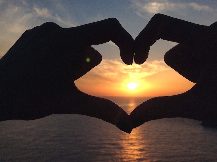 Hands held in heart shape around the setting sun of Vatolakkos, Greece