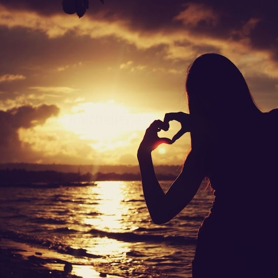 Woman holds hands up in a heart shape during sunset overlooking water