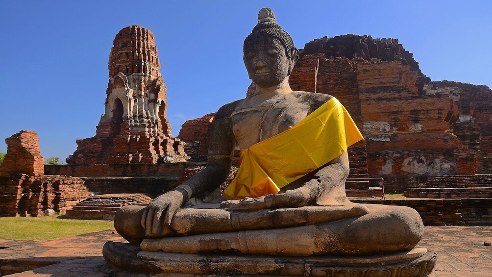 Gigantic Buddha statue with yellow sash sitting in front of ancient temples