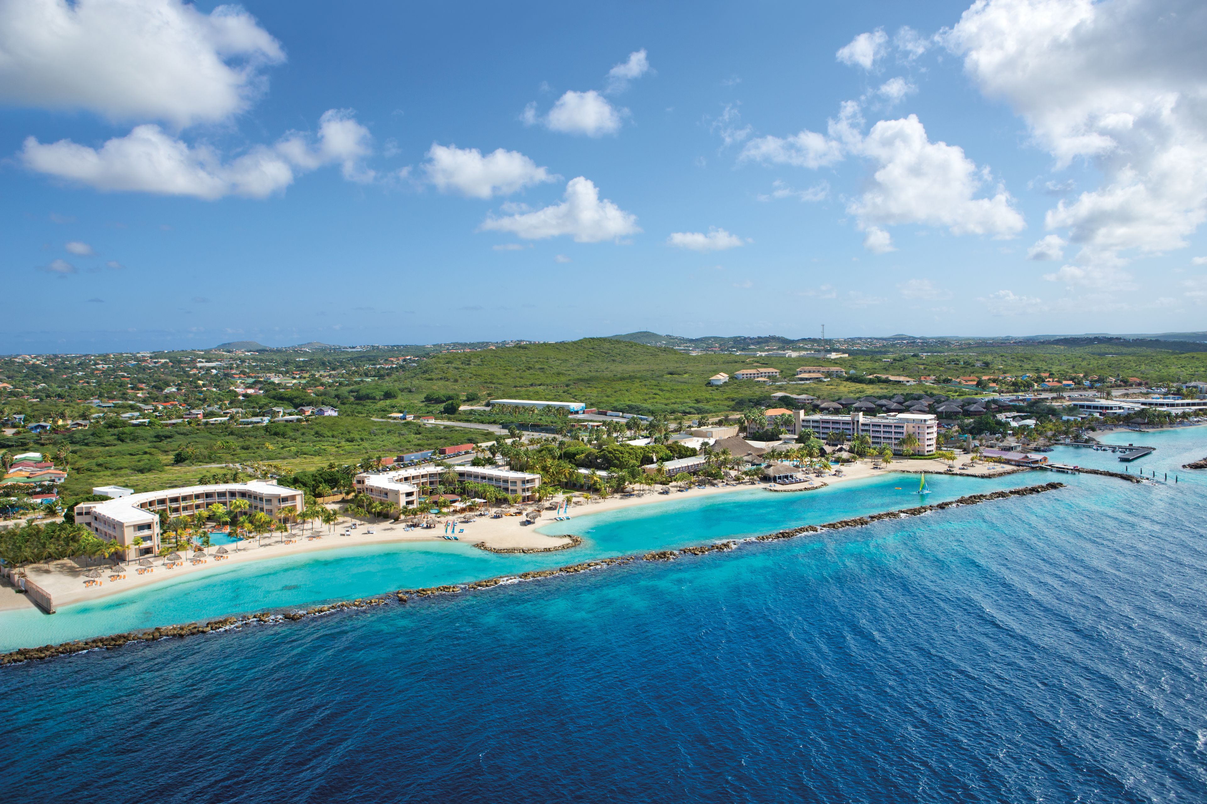 An aerial view of a coastal resort with multiple buildings, a sandy beach, and clear turquoise waters.