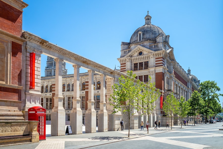 Daytime view of the Victoria and Albert Museum in London, showcasing its grand Victorian architecture, domed roof, and columned entrance under a clear blue sky.