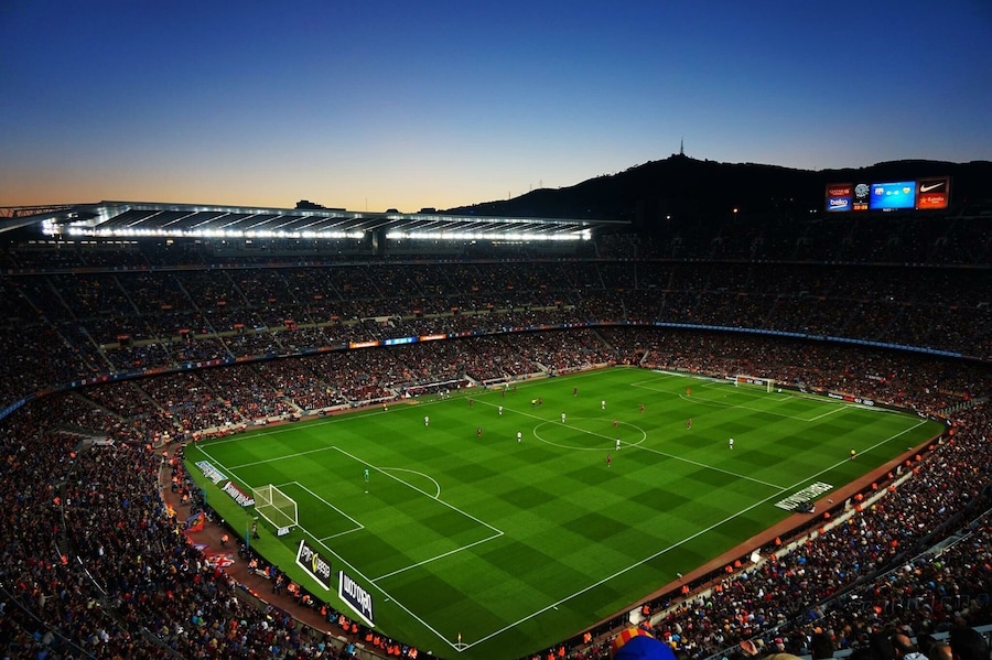 Evening football match at Camp Nou stadium in Barcelona, with a packed crowd and city hills visible at sunset.
